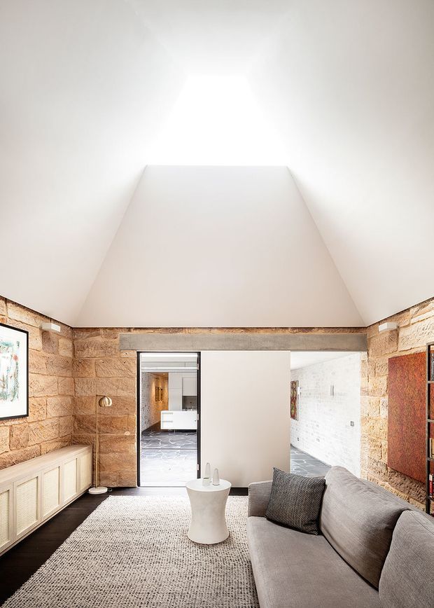 A refined living room in a renovated stone-and-brick interior, featuring a large grey sofa, textured light rug, pale timber sideboard, white sculptural side table, and dark flooring. The room has exposed warm sandstone walls, a dramatic vaulted white ceiling, and wide openings leading to a contemporary kitchen or adjoining space.