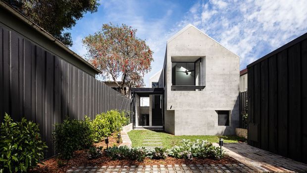 A compact contemporary residential exterior viewed from a landscaped front approach. The home features a simple gabled concrete volume with a large recessed window, dark-framed glazing, and a dark timber or metal boundary fence. A narrow garden path, lawn strips, low planting, and brick-paved foreground lead toward the entrance.