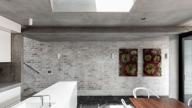 Contemporary open-plan kitchen and dining area with a white island, dark stone flooring, exposed pale brick wall, concrete ceiling with a large skylight, and black-framed glazing. A timber dining table and floral artwork add warmth to the otherwise restrained monochrome interior.