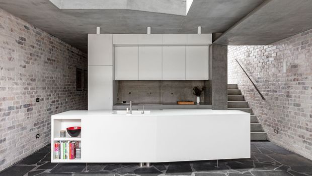 Minimalist open-plan kitchen with a large white island and integrated cabinetry, set within a raw industrial shell of exposed brick walls, concrete ceiling, concrete structural column, and dark stone flooring. A stairway rises beside the kitchen, and a rooflight brings natural light into the space.