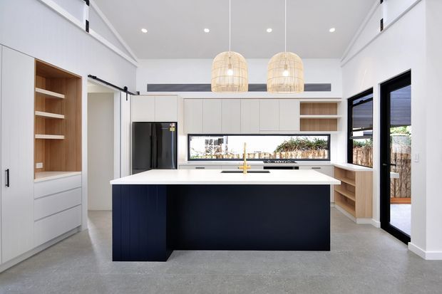 Bright contemporary kitchen with a large dark island and white stone benchtop, pale integrated cabinetry, timber open shelving, black-framed glazing and two woven pendant lights. The room has a polished concrete-look floor and a high pitched ceiling with exposed structural-style beams.