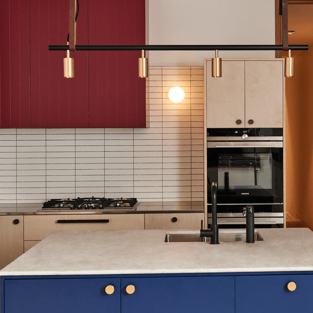 Contemporary kitchen featuring a large pale stone island with integrated sink and dark tapware, deep blue lower cabinetry with brass knobs, light timber cabinetry, a black gas cooktop, stacked oven appliances, and a white horizontally tiled splashback. A burgundy red wall panel and linear brass pendant lighting add bold colour and contrast.