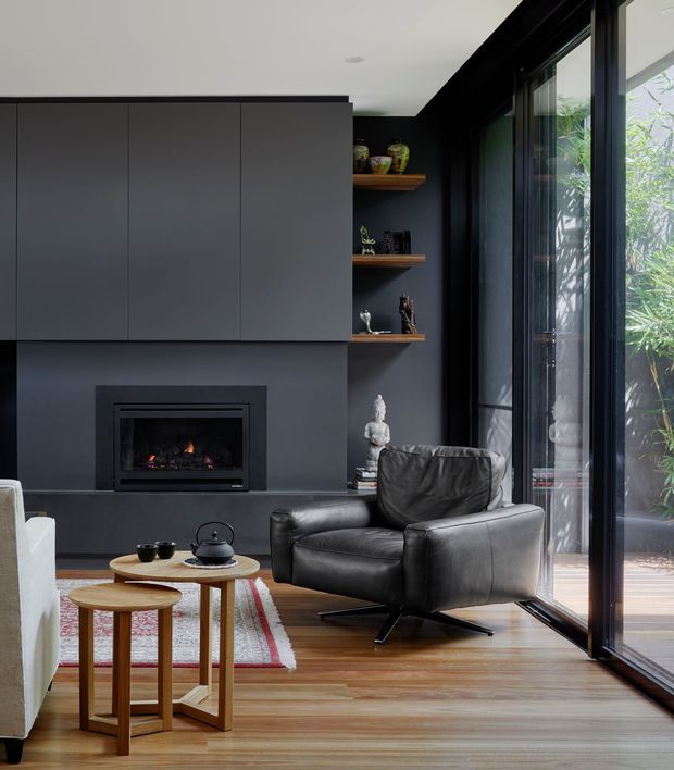 A refined contemporary living room with a dark built-in fireplace wall and concealed cabinetry, open timber shelving, a black leather lounge chair, nesting timber coffee tables, and a pale sofa edge. Full-height black-framed glazing opens to greenery and a timber deck, while warm timber flooring and a patterned rug soften the dark palette.