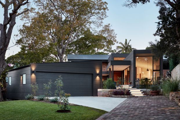 A contemporary single-storey home viewed from the front, featuring dark horizontal cladding, a broad integrated garage, a recessed timber-accented entry, large warm-lit glazing, concrete steps, and a brick-paved driveway. Mature trees and landscaped lawn frame the facade.