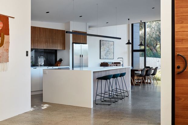 Open-plan contemporary kitchen and dining area viewed from an entryway. The space features a large white island with teal bar stools, warm timber cabinetry, dark stone or tile splashback, polished concrete flooring, pendant lighting, and full-height windows overlooking greenery. A timber entry door and artwork are visible at the edges of the frame.