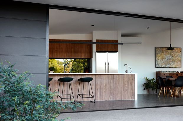 Contemporary open-plan kitchen and dining area viewed from an exterior or semi-enclosed threshold. The space features a timber-clad kitchen island with two black bar stools, dark flooring, white walls and ceiling, large glazing, a timber roller blind, pendant lighting, and a dining table beyond. Dark horizontal exterior cladding frames the left side, while indoor planting and artwork add warmth.