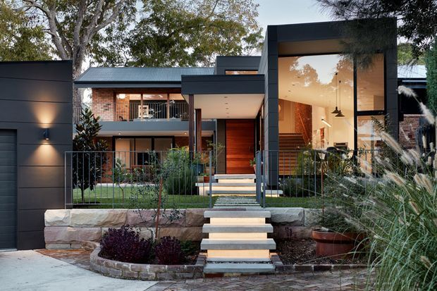 Exterior view of a contemporary multi-level residence with dark charcoal cladding, warm timber entry door and screening, extensive floor-to-ceiling glazing, and a stepped entry path through landscaped garden beds. Stone retaining walls, exposed brickwork, metal roofing, and a sheltered balcony contribute to the modern residential facade.