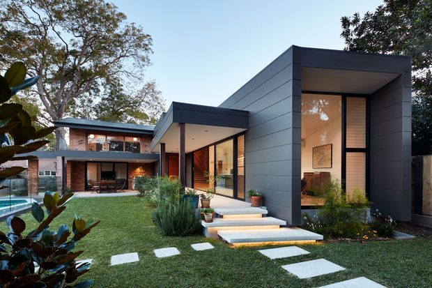 Contemporary two-storey residence with a dark charcoal, flat-roofed extension and broad horizontal cladding, connected to a lighter brick-and-glass rear volume. Large glazed openings, timber entry door, covered outdoor entertaining area, landscaped lawn, stepping-stone path, and a swimming pool are visible.