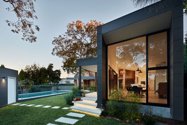 Contemporary single-storey house exterior at dusk, featuring dark vertical cladding, expansive floor-to-ceiling glazing, a projecting flat-roofed volume, and illuminated open-plan interiors visible through the windows. A landscaped lawn with stepping stones, planted borders, and a swimming pool forms the foreground and side yard.