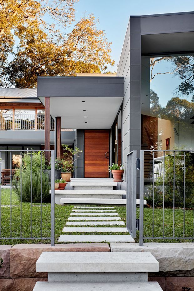 Contemporary residential house viewed from the street, featuring a charcoal grey rectilinear facade, large framed glazing, a covered entry porch, warm timber front door, exposed timber posts, concrete steps and stepping-stone path, and a slim metal front fence. Mature deciduous trees and landscaped planting surround the entrance.