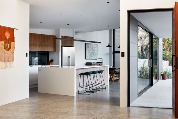 Open-plan contemporary kitchen and entry area with a large white island, integrated seating for four, dark timber cabinetry, stainless-steel appliances, polished concrete flooring, and black-framed glazing. A glazed entry passage opens to a planted courtyard, while a linear suspended light and minimalist furnishings reinforce the clean modern aesthetic.