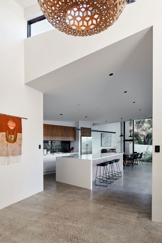 A bright, open-plan contemporary interior viewed from a high-ceilinged entry or living area toward a white kitchen and dining space. The kitchen features a large central island with waterfall-style white surfaces, integrated appliances, warm timber cabinetry, dark stools, and black pendant lighting. Polished concrete flooring, expansive glazing, and a sculptural woven timber pendant add texture and visual interest. A dining table and chairs are visible beyond the island, with greenery outside the windows.