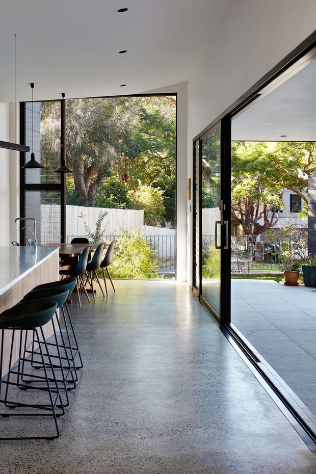 A bright, contemporary open-plan kitchen and dining area with a long white island, black bar stools, pendant lights, and a timber dining table. Full-height black-framed glazing and large sliding doors open directly to a paved outdoor courtyard and garden, creating a strong indoor-outdoor connection. The room has a polished grey concrete floor, white walls and ceiling, and abundant natural light.