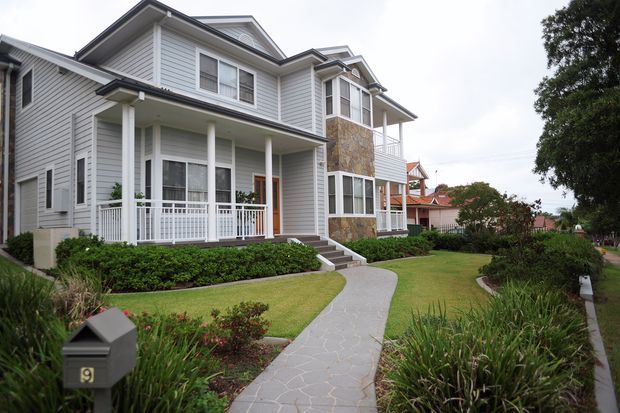 Exterior view of a large multi-storey residential house with light grey horizontal weatherboard cladding, white trim, covered verandas and balconies, stone-clad facade sections, and a landscaped front garden with a curved paved walkway.
