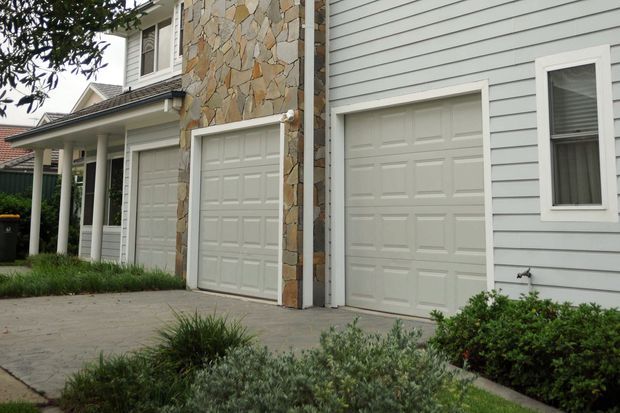 Exterior view of a residential facade with two white paneled garage doors, pale horizontal weatherboard cladding, a central irregular natural-stone feature wall, white-trimmed windows, and a paved driveway bordered by low landscaping.
