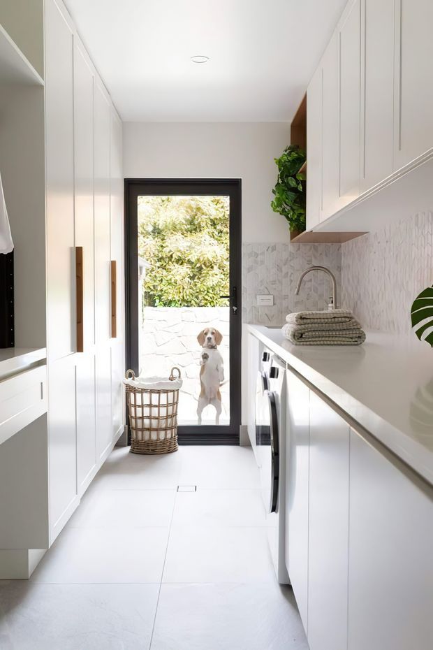 A bright, narrow contemporary laundry room with floor-to-ceiling white cabinetry on both sides, a long white benchtop, integrated appliances, a curved chrome tap and pale geometric tiled splashback. A black-framed glazed door opens to an outdoor area, with a woven laundry basket and folded towels adding texture. Natural greenery is visible outside and through an open shelf, while a dog stands beyond the door.