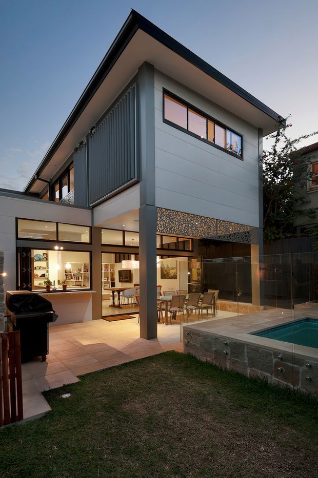 A dusk exterior view of a contemporary multi-level residence with a sharp pitched roof, pale horizontal-clad walls, dark window frames, and extensive floor-to-ceiling glazing. The house opens onto a tiled outdoor entertaining area with dining furniture, a swimming pool, lawn, and an integrated barbecue zone. A perforated decorative screen and vertical privacy louvres add architectural detail to the facade.