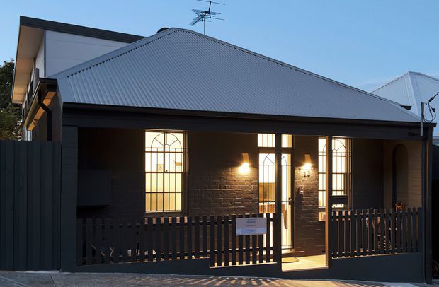 Street-facing exterior of a compact single-storey dwelling with a steep hipped corrugated metal roof, dark painted brick walls, and a covered front porch enclosed by a dark vertical timber or metal picket fence. Tall arched-top windows and glazed entry doors feature decorative muntins, with warm interior and wall lighting visible at dusk. A contemporary dark addition and neighbouring roofline are visible behind.
