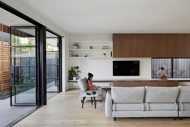 Bright contemporary living room with a large light fabric sectional sofa, pale timber flooring, white walls, open white shelving, and a built-in media wall with dark timber cabinetry. Full-height black-framed glazing opens toward an outdoor area with timber screening. The room has a clean, refined and inviting appearance.