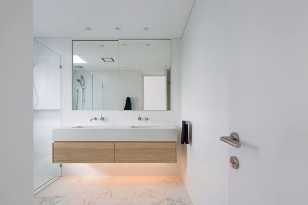 Bright minimalist bathroom viewed from the doorway, featuring a floating double vanity with pale timber drawers, white countertop and twin basins, a large mirrored cabinet, frameless glass shower enclosure, recessed ceiling lights, and pale marble-look floor tiles. A partially open white door with brushed metal hardware is visible in the foreground.
