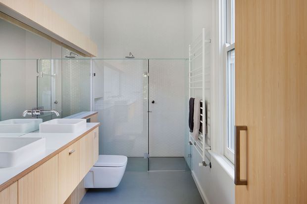 Bright, contemporary bathroom with a long light-wood vanity featuring multiple white vessel basins, a wall-hung toilet, and a large frameless glass shower enclosure. Pale walls, grey floor tiles, white heated towel rail, and tall windows create an airy, functional space. A timber door and matching vertical timber cabinetry add warmth.