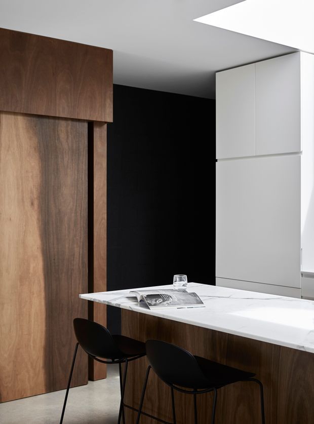 A refined contemporary kitchen featuring dark timber cabinetry, a black feature wall, tall white integrated cabinetry, and a white marble-look island with seating for two. A large rectangular skylight or ceiling opening provides natural light, creating a high-contrast, minimalist composition.
