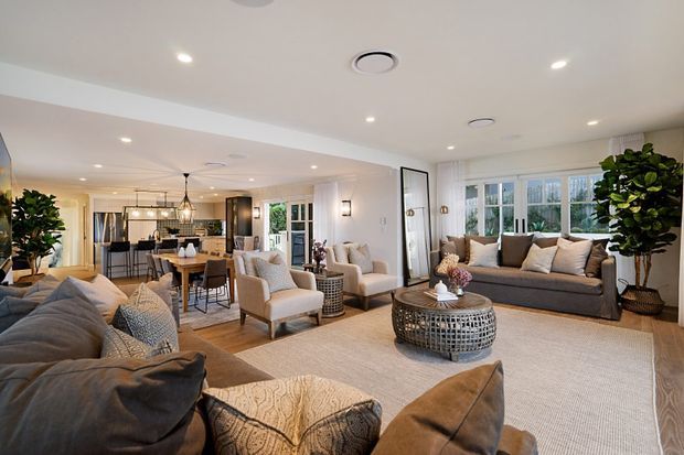 A spacious, bright open-plan living room with a large sectional sofa, upholstered armchairs, a woven area rug, and a round textured coffee table. The room opens directly into a dining and kitchen area, with extensive windows, recessed lighting, indoor plants, and warm neutral furnishings.