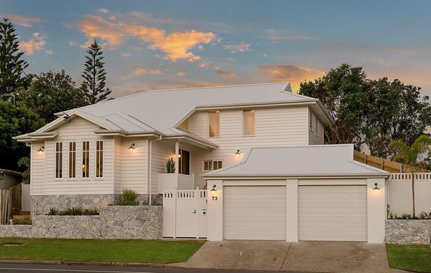 Street-facing exterior view of a refined single-storey white weatherboard home with a broad pitched metal roof, projecting gables, tall multi-pane windows, a stone retaining wall, and an attached double garage. The home is set behind a low landscaped frontage with a paved driveway and white boundary fencing, photographed at dusk.