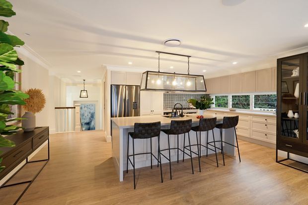 Bright, spacious open-plan kitchen with a large central island, four black bar stools, stainless-steel refrigerator, pale cabinetry, tiled backsplash, timber-look flooring, and a large linear pendant light. A dining or living area and hallway are visible beyond the kitchen, with dark built-in shelving and indoor plants adding contrast.