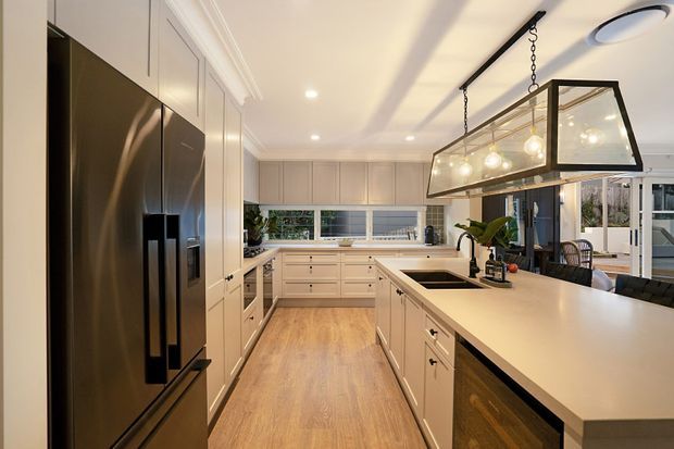 A bright, spacious contemporary kitchen with a long central island, pale cabinetry, light timber flooring, a dark integrated refrigerator, and a large rectangular glass-and-metal pendant light. The kitchen opens toward a glazed outdoor area and includes a wide horizontal window above the rear benchtop.