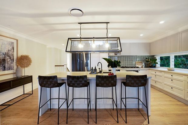 A bright, spacious kitchen with a large central island and four dark woven bar stools. The room features pale cream cabinetry, white benchtops, a grey-green tiled splashback, timber flooring, stainless-steel appliances, and a prominent rectangular pendant light over the island. A sideboard and artwork sit along the adjacent wall, with greenery visible through the windows.