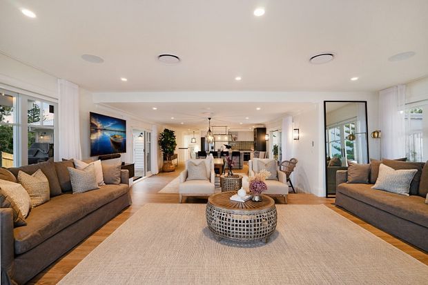 A bright, open-plan living room with two large neutral sofas facing inward across a broad natural-fibre rug, a pair of light upholstered armchairs, and a round woven coffee table. White walls, recessed ceiling lights, large windows with sheer curtains, a tall black-framed mirror, timber flooring, and a visible dining and kitchen area create an airy, refined residential interior.