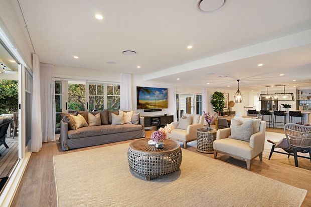 Bright open-plan living room with a large neutral rug, grey sofa, cream armchairs, woven round coffee table, timber flooring, and soft white walls. Sliding glass doors and large windows open toward greenery, while a dining area and kitchen are visible in the background. Recessed ceiling lighting and pendant lights create an airy, relaxed interior.