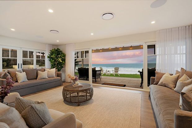 Bright, spacious coastal living room with large sofas and armchairs arranged around a round woven coffee table and a large neutral rug. Full-height glazed doors and windows open to a deck overlooking the ocean, while sheer curtains, recessed lighting, and abundant neutral upholstery create a relaxed beachside atmosphere.
