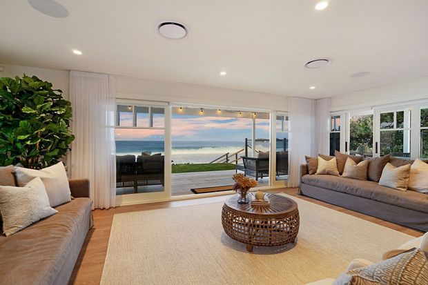 Bright coastal living room with warm timber flooring, neutral upholstered sofas, a large woven rug, and a round woven coffee table. Full-height glazing and sliding doors open toward a deck with direct ocean views, while sheer curtains and recessed lighting create an airy, relaxed interior.