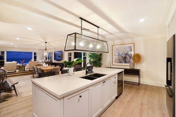 Bright open-plan kitchen and dining area with a large white island, integrated sink and dark cooktop, framed cabinetry, timber-look flooring, and a large rectangular pendant light. A dining table, living area, artwork, plants, and expansive windows are visible in the background.