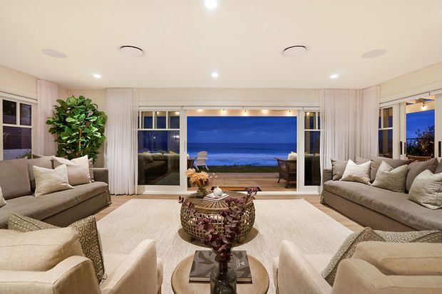 Elegant coastal living room with symmetrical seating arranged around a central coffee table, large retractable or folding glazed doors opening to an ocean view, sheer curtains, neutral upholstered sofas and armchairs, a pale textured rug, and a sculptural woven coffee table. The room has a bright, refined atmosphere with recessed ceiling lighting and abundant natural light.