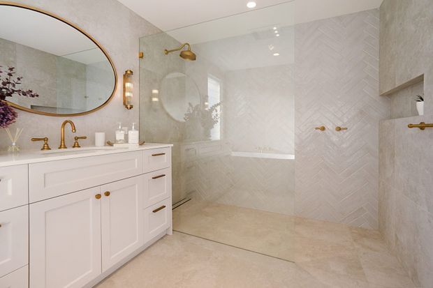 Bright, refined bathroom with a large white shaker-style vanity, brushed brass plumbing fixtures and hardware, an oversized rounded-rectangle mirror, and a frameless glass shower enclosure. The shower and walls are finished in pale herringbone-pattern tile, with a built-in niche and matching brass fittings. The palette is warm white, cream, and soft beige with brass accents.