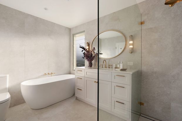 Bright, refined bathroom featuring a freestanding white bathtub, wall-to-wall pale stone-look tiles, a white built-in vanity with brass hardware, round framed mirror, wall sconces, and a frameless glass shower enclosure.