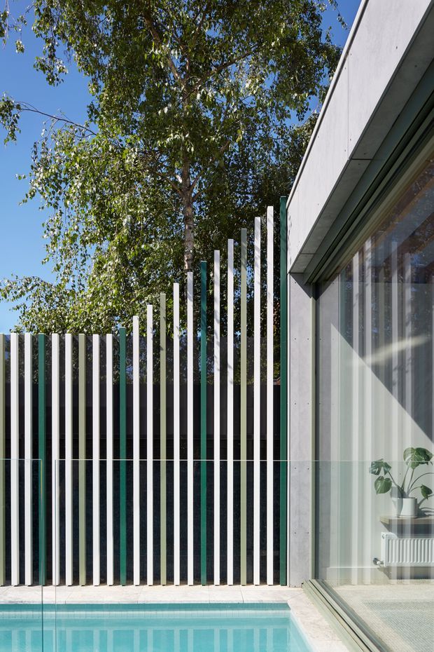 Exterior architectural view of a contemporary building beside a swimming pool. A tall sculptural screen of evenly spaced vertical white fins rises in front of dark green privacy glazing and steps upward toward the building. The facade features pale concrete or fibre-cement panels, extensive floor-to-ceiling glazing, and a visible interior with a potted plant and radiator. Bright blue sky, mature trees, and turquoise pool water frame the scene.