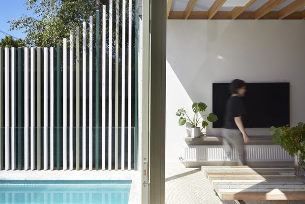 A bright contemporary interior opens directly onto a swimming pool through large glazed doors. The living area features a wall-mounted television, low built-in white joinery, pale textured flooring, indoor plants, and a striking exposed timber beam ceiling. Outside, a tall vertical slatted privacy screen borders the pool.