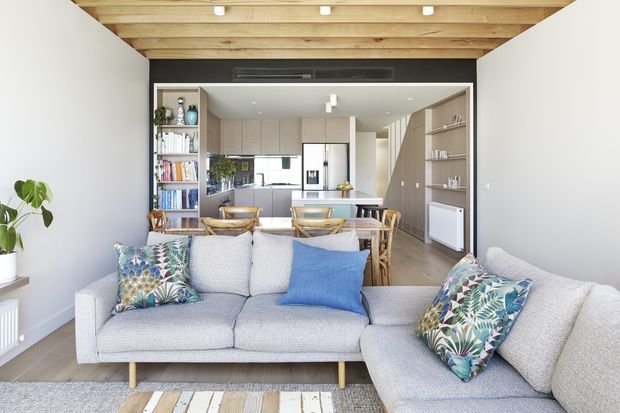 Bright open-plan living space with a large light-grey sectional sofa in the foreground, timber flooring, pale walls, and a striking exposed timber ceiling with beams. A dining area and contemporary kitchen with light cabinetry are visible beyond, with open shelving, indoor plants, and patterned blue-green cushions adding colour.