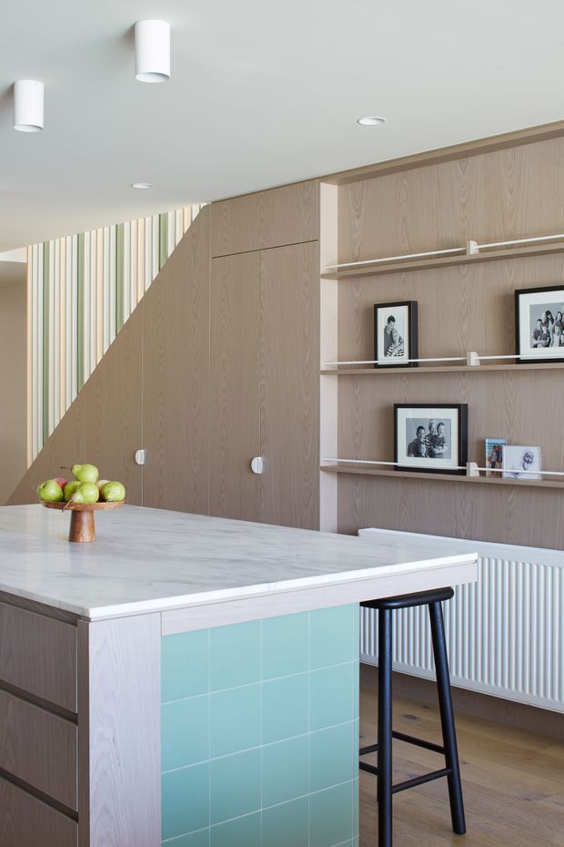 A bright, contemporary kitchen centred on a large pale stone island with a turquoise tiled end panel and a black bar stool. The room features extensive light timber wall panelling and built-in cabinetry, open display shelves with framed photographs, a white panel radiator, recessed and cylindrical ceiling lights, and a partially screened stair or upper-level area with vertical pastel-toned slats. The palette is light timber, white, pale grey, and aqua, creating a clean, refined interior.