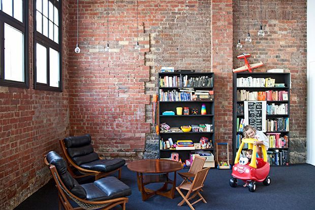 A loft-style interior with exposed red brick walls, tall dark-framed multi-pane windows, and a dark carpeted floor. Two black leather lounge chairs and a round timber coffee table form a seating area, while open bookshelves filled with books and toys line the rear wall. Small pendant lights hang across the room, and a child's ride-on toy is visible near the shelving.