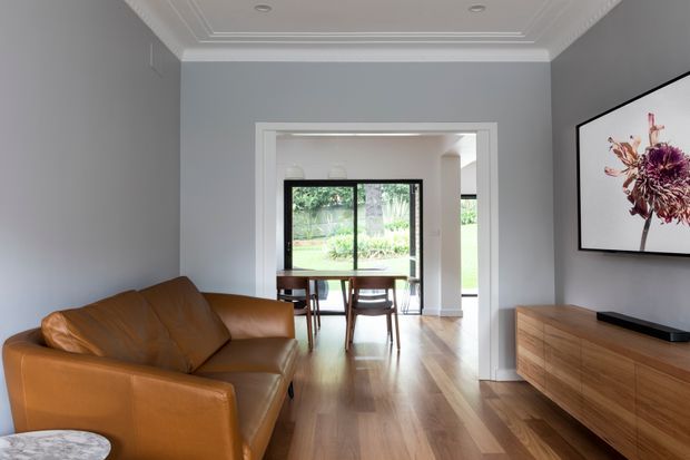 A refined open-plan living room with a large tan leather sofa, timber flooring, a low timber media console, and a wall-mounted television displaying floral artwork. The room opens through a wide trimmed doorway to a dining area with a timber table and chairs, with garden views beyond. Pale grey walls, white decorative ceiling cornices and recessed lighting create a calm, contemporary interior.