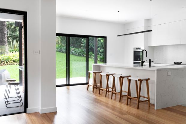 Bright, contemporary open-plan kitchen with a large white island and waterfall stone-look benchtop, integrated appliances, black tapware, and seating for several people on timber stools. White full-height cabinetry and a minimalist linear pendant light are paired with warm timber flooring. Large black-framed glazed doors and windows open toward a leafy garden and lawn.