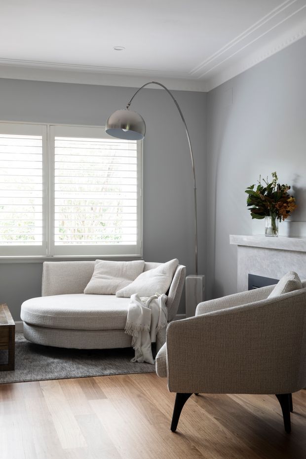 Refined living room with soft grey walls, ornate ceiling cornices, white plantation shutters, pale timber flooring and a light upholstered curved chaise-style sofa. A sculptural arched floor lamp, textured armchair, neutral rug, fireplace surround and floral arrangement create a calm, elegant interior.