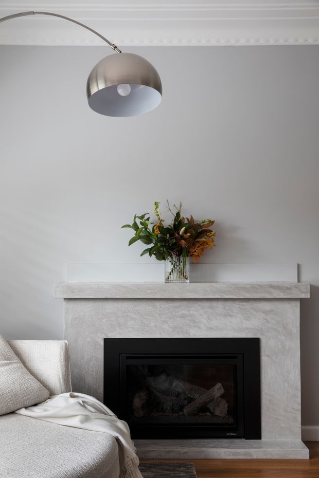 A refined living room vignette with a textured pale fireplace surround and mantel, black built-in fireplace insert, neutral upholstered sofa with a draped throw, and a vase of foliage and flowers. A curved-arm metallic pendant light hangs beneath decorative ceiling cornice detailing. The palette is soft grey, white, black, and warm timber.
