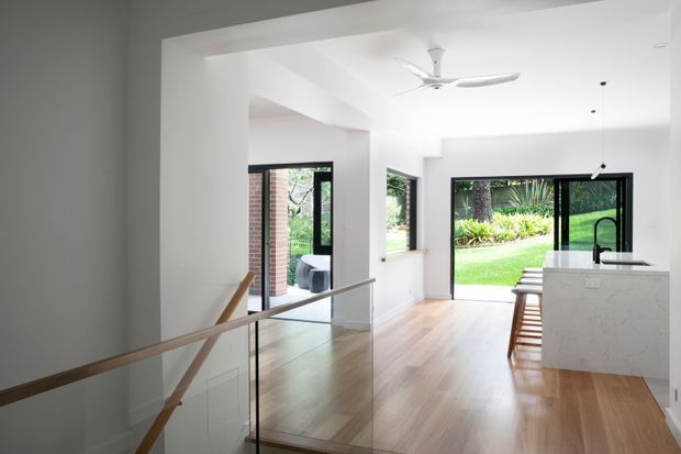 Bright contemporary open-plan interior viewed from a stair landing, with white walls and ceiling, pale timber flooring, black-framed glazing opening to a green garden, a glass balustrade with timber handrail, and a white stone kitchen island with seating. A ceiling fan and pendant lights complete the clean, airy space.