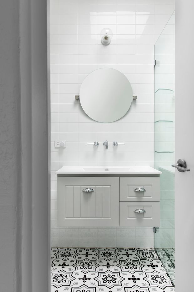 A compact, bright bathroom viewed through an open doorway. The room features white horizontal wall tiles, a floating white vanity with chrome handles and taps, a round frameless mirror, a glass shower enclosure, and highly decorative black-and-white patterned floor tiles. The palette is predominantly white and grey with strong monochrome contrast.